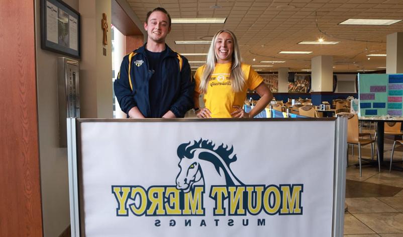 Two students posing in front of a Mount Mercy banner